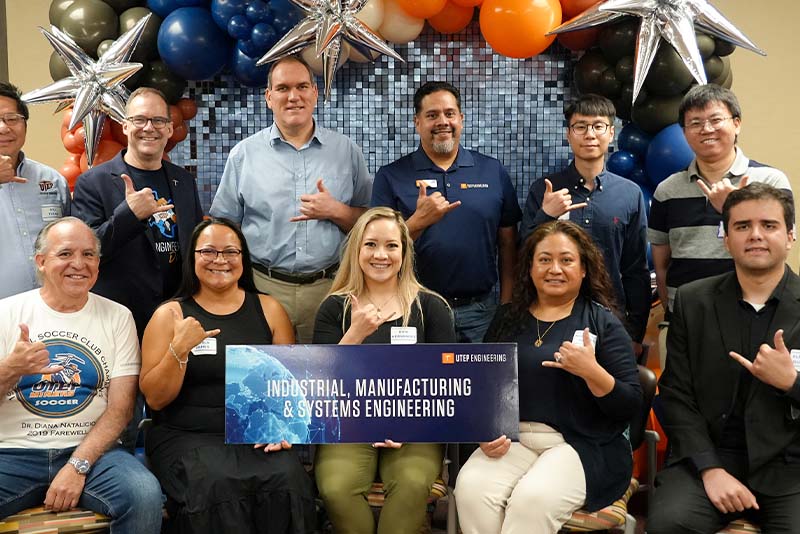 Group of attendees pose together during a UTEP Engineering homecoming gathering while holding a large “Computer Science” banner, seated and standing in front of a backdrop decorated with orange, blue, black, and silver balloons while making hand gesture signs for the departmental photo.